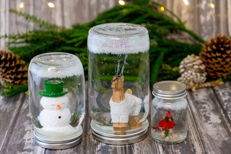 Three DIY snow globes with reindeer, snowman, and cardinal inside jars on table.