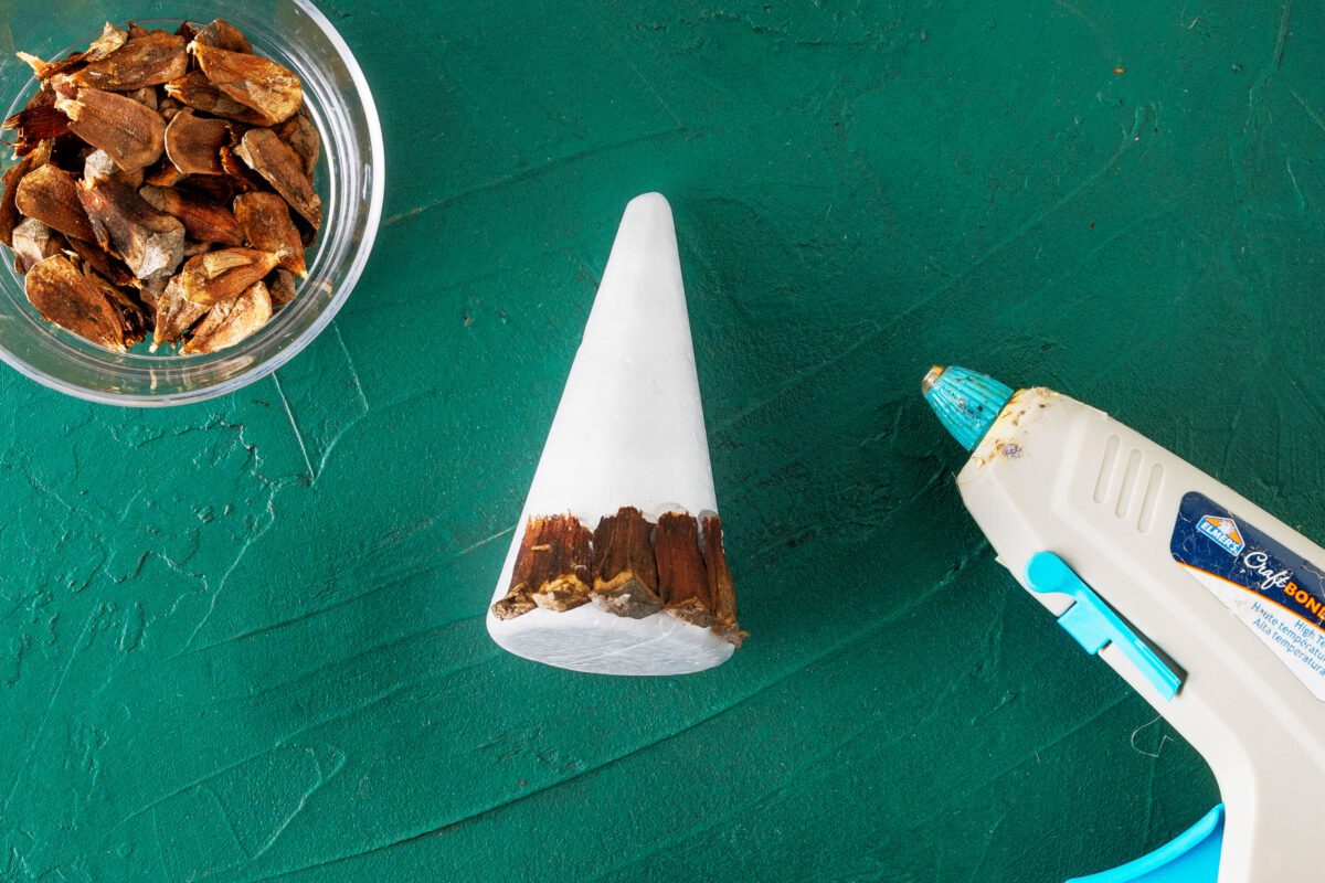 Bowl of pine cone scales, cone base, and glue gun on green background.