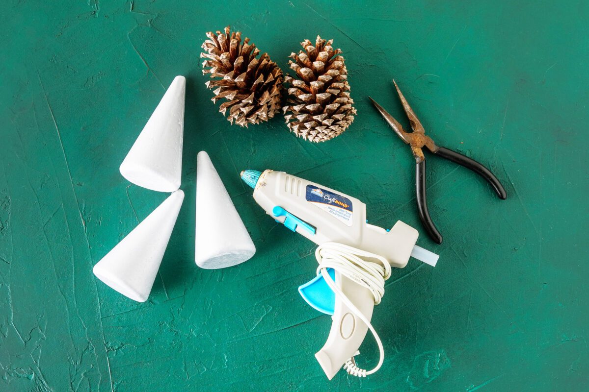 Crafting materials including glue gun, foam cones, and pine cones on green surface.