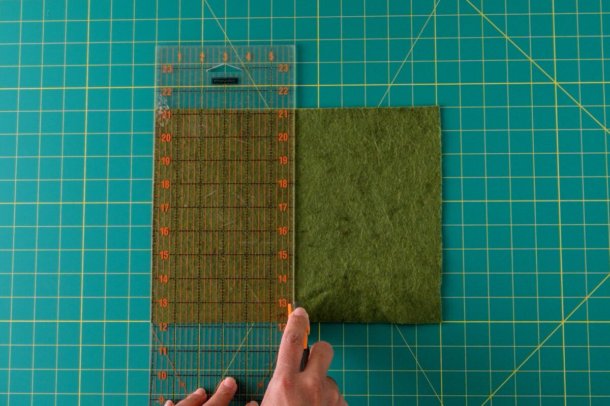 Hand cutting green felt piece using a transparent ruler and rotary cutter.