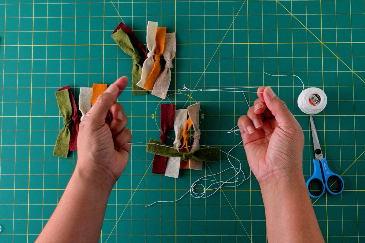 Stringing colorful felt bows with thread and scissors on a gridded mat.