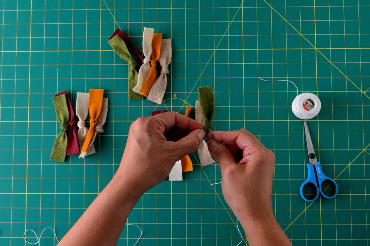 Tying green felt bow using thread and needle with colorful bows nearby.